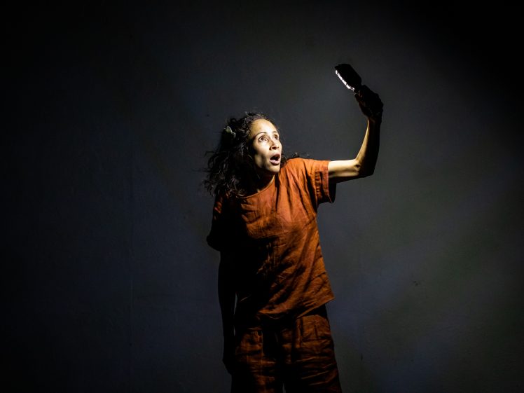 Image of A person wearing an orange outfit stands in a dark space, illuminated only by the light from a mobile or small device held in their outstretched hand, with a surprised expression on their face.