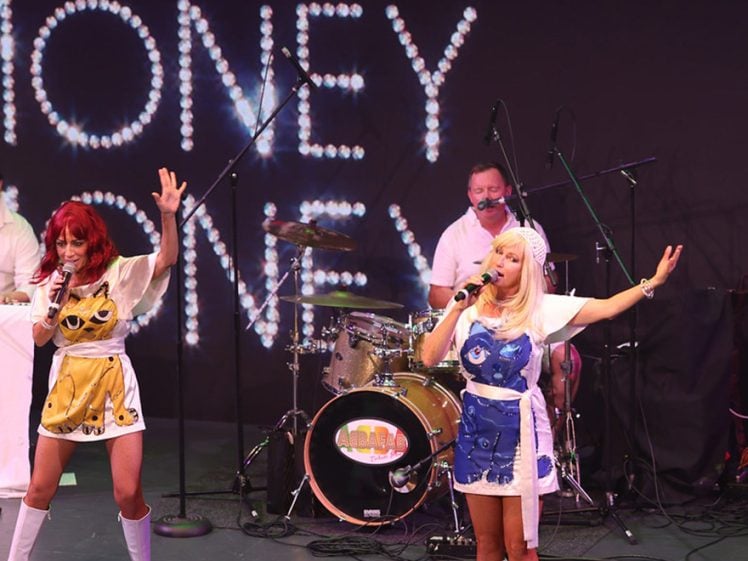Image of Two women in colourful, retro costumes sing on stage with microphones, while a drummer plays in the background. Large “MONEY MONEY MONEY” lights up the backdrop, creating a lively concert atmosphere.
