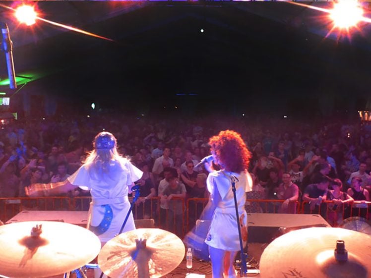 Image of View from behind a drum kit on stage, two performers in white costumes sing to a large, energetic crowd under bright stage lights in a dark indoor venue.