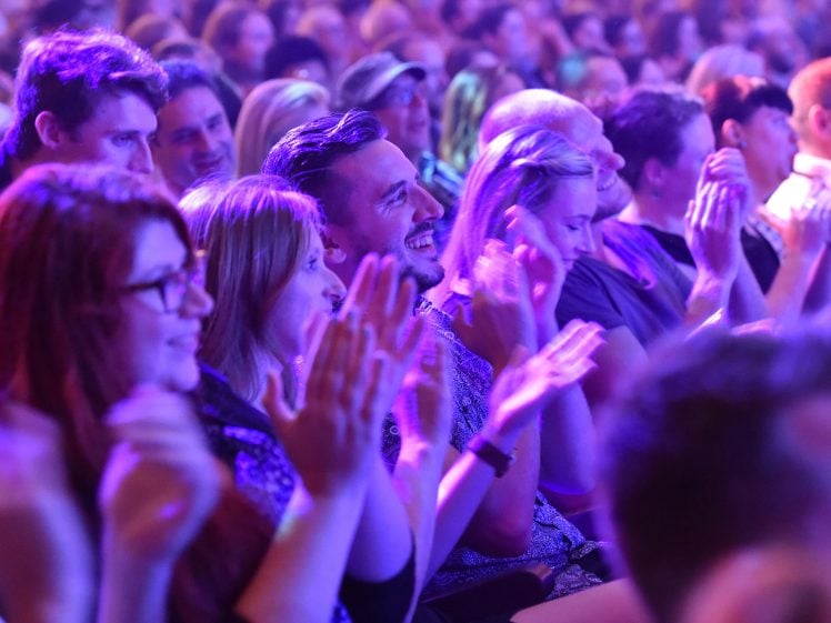 Image of An audience sits closely together in a brightly lit venue, smiling and clapping enthusiastically. The lighting casts colourful purple and blue tones across the crowd, highlighting their joyful expressions.