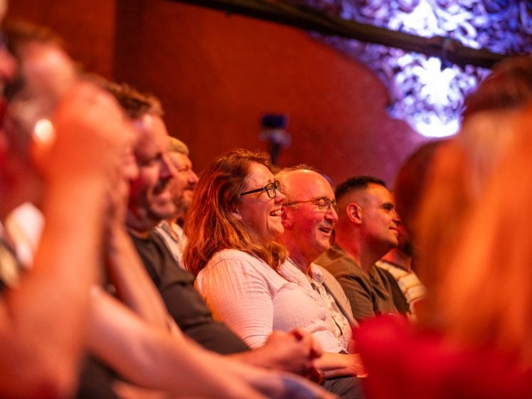 Image of A group of people in a dimly lit room, with a woman in the foreground laughing. The background has red and purple lighting.