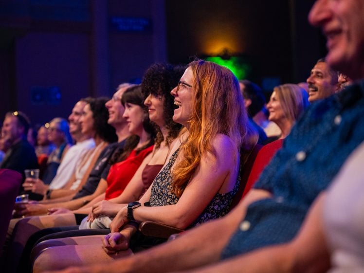 Image of A group of people sit in theatre seats, smiling and laughing as they watch a live performance. The focus is on a woman with long red hair in the centre. The audience appears to be enjoying the show.