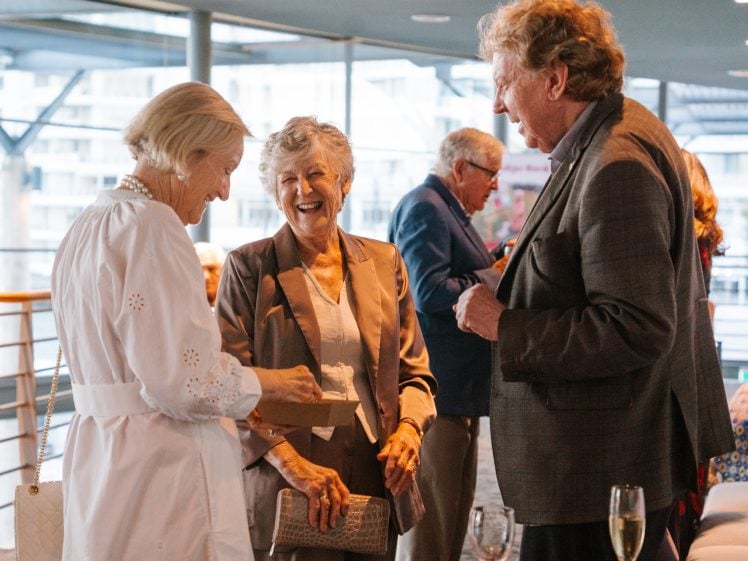 Image of Three older adults—two women and one man—stand indoors, smiling and chatting. They are dressed in semi-formal attire, with drinks on a nearby table, suggesting a social or celebratory event.