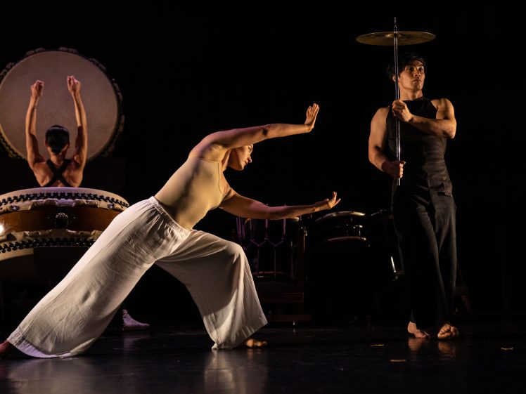 Image of Three performers on stage: one plays a large drum in the background, one in white trousers strikes a dramatic pose in the centre, and one in black holds a spinning plate on a stick to the right. The stage is dimly lit.
