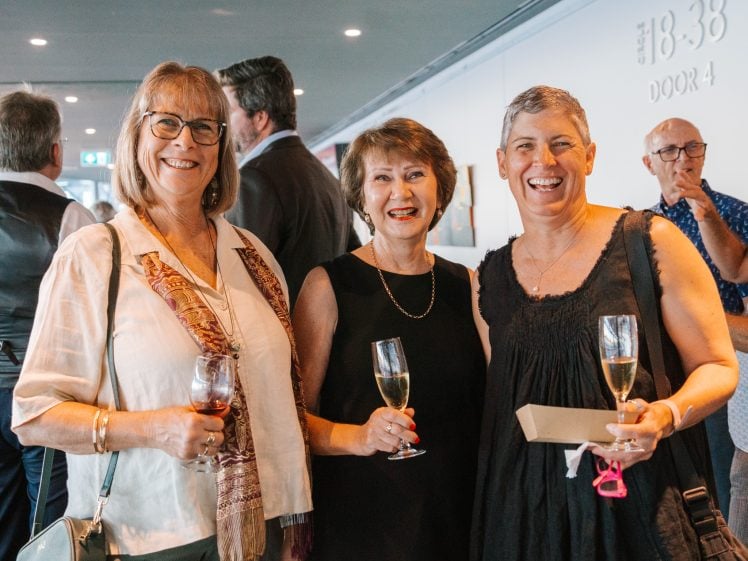 Image of Three women stand together indoors, smiling and holding drinks at a social event. Other people are mingling in the background. The women are dressed in semi-formal clothing and appear happy and engaged.