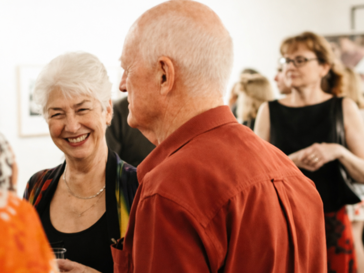 Image of A group of people socialise at an indoor event; an older woman with white hair smiles whilst talking to an older man in a red shirt, with others mingling in the background.