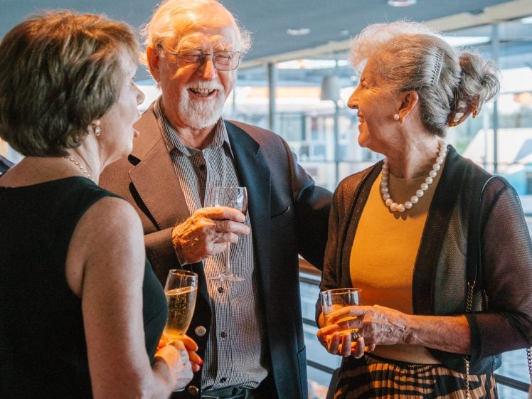 Image of Three older adults dressed in formal attire stand together indoors, smiling and holding drinks whilst engaging in conversation at what appears to be a social event or gathering.