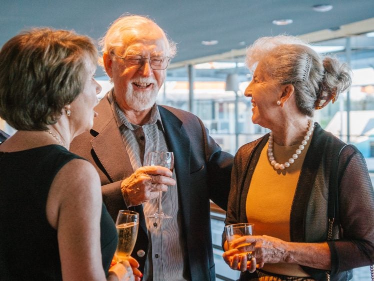 Image of Three older adults dressed in formal attire stand together indoors, smiling and holding drinks whilst engaging in conversation at what appears to be a social event or gathering.