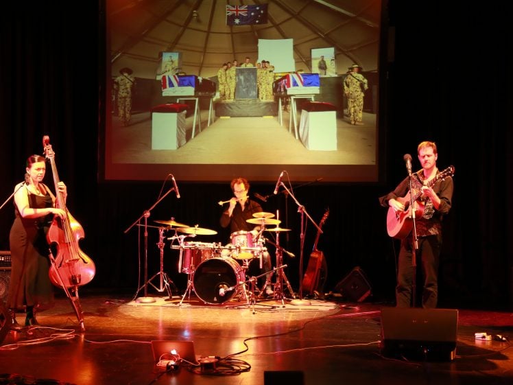 Image of Three musicians perform on stage with a double bass, drums, and guitar. Behind them, a large screen displays an image of soldiers standing in front of a military memorial with flags. Microphones and instruments are on stage.