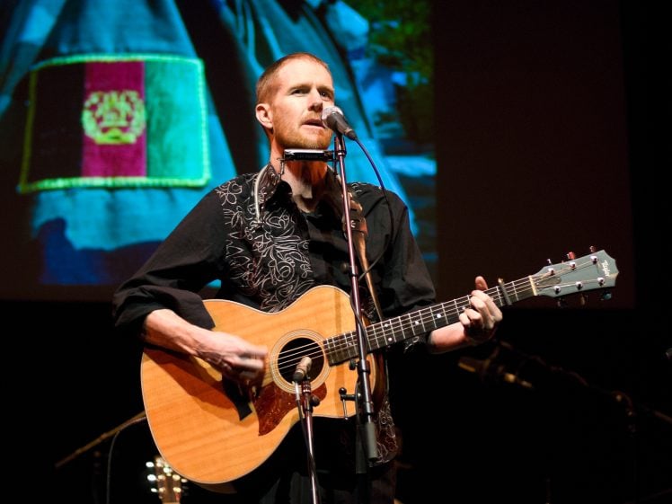 Image of A man with short hair and a reddish beard plays an acoustic guitar and sings into a microphone on stage. He is wearing a black shirt with white embroidery. A colourful image is projected in the background.