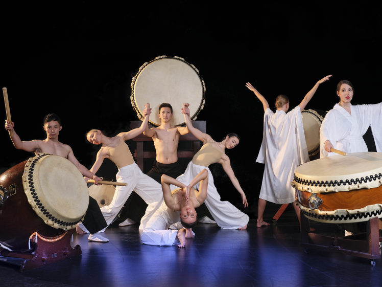Image of Six performers are on stage with traditional Japanese taiko drums, posing dynamically. Some are in white costumes, others shirtless, and one is upside down, with a dark background emphasising their dramatic expressions and movements.