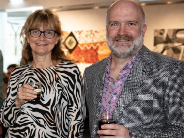Image of A woman in a zebra-print top and glasses stands next to a man in a grey blazer and pink shirt. Both are smiling and holding drinks at an indoor event with patterned artwork on the wall behind them.
