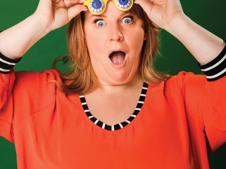 Image of A surprised woman with light brown hair holds yellow novelty glasses with blue eyeballs over her eyes. She wears an orange top with black and white striped trim against a green background.