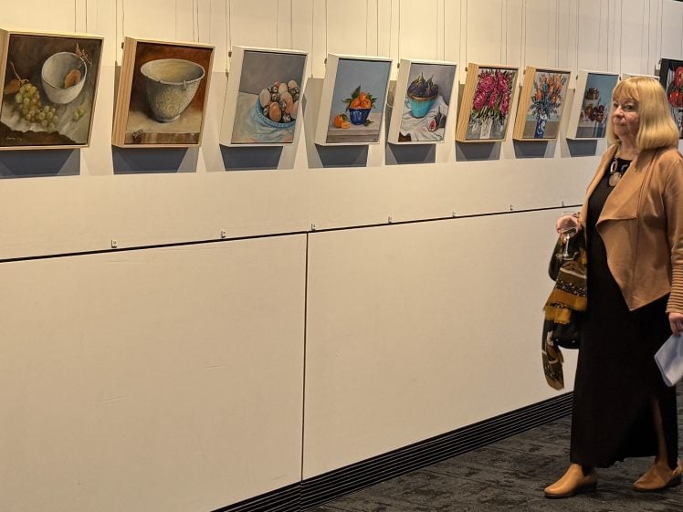 Image of A woman holding a wine glass and a bag walks past a wall displaying several colourful paintings, including still lifes and flowers, in an art gallery setting.