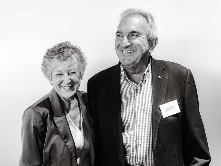 Image of An older woman and man stand smiling together against a plain backdrop. Both wear business attire with name badges; the woman has short hair and the man has grey hair. The image is in black and white.