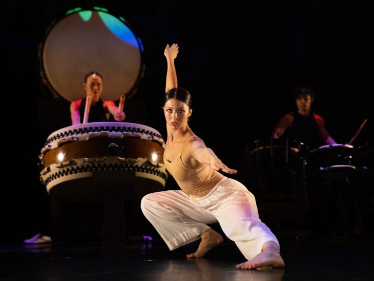 Image of A dancer in white trousers and a beige top poses gracefully on stage, while two musicians play large traditional drums in the background under dramatic lighting.