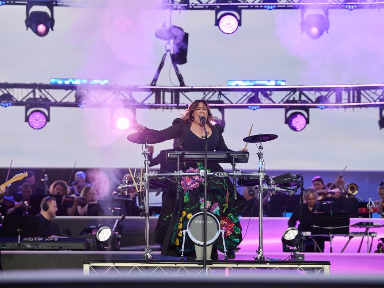 Image of A musician stands on stage singing passionately whilst playing electronic drums, surrounded by purple lights and a live orchestra in the background during an outdoor concert.