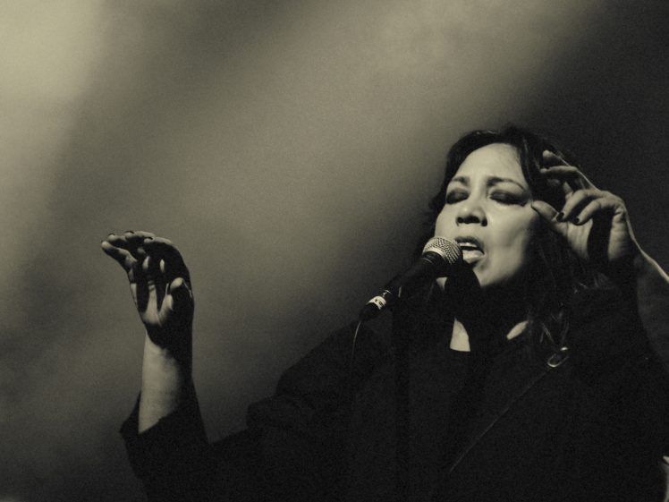 Image of A woman sings passionately into a microphone on stage, eyes closed and one hand raised, with dramatic lighting and a smoky background in a black-and-white photo.