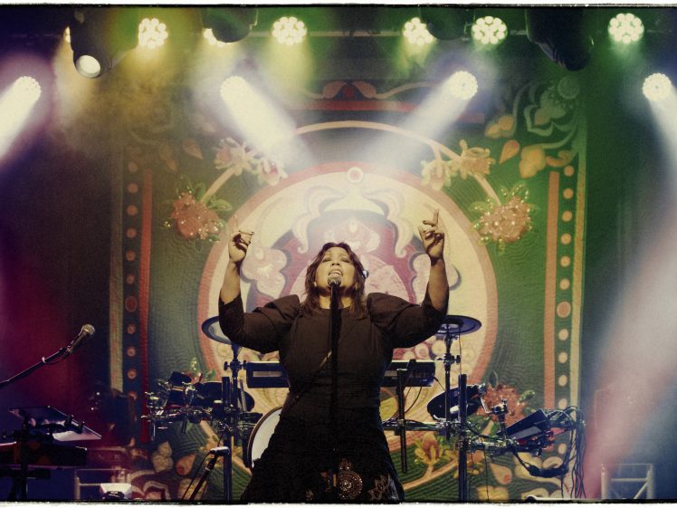 Image of A singer performs passionately on stage under colourful lights, with arms raised. A vibrant, ornate backdrop with circular patterns and floral designs is visible behind, and musical equipment surrounds the performer.