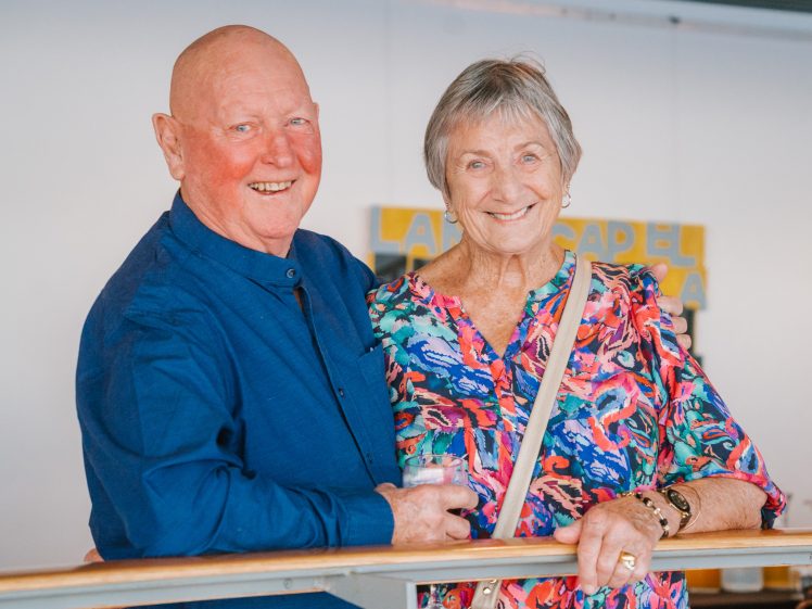 Image of An elderly man in a blue shirt and an elderly woman in a colourful blouse smile together whilst standing by a railing indoors.