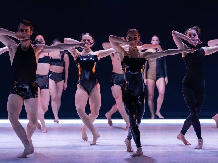 Image of A group of dancers in black and beige costumes perform on stage, striking dynamic poses with one arm bent in front of their faces, under dramatic lighting.