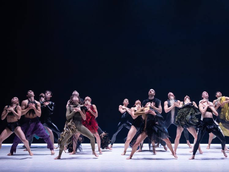 Image of A group of dancers performs on stage, wearing varied, flowing costumes in dark and earthy tones, striking expressive poses with arms crossed over their chests against a dark background.