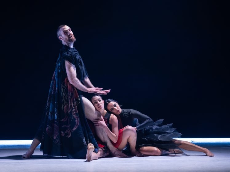 Image of Three ballet dancers pose dramatically on stage; one stands with a black cape, while two others in black and red costumes sit and lean against him, all illuminated by soft stage lighting against a dark background.