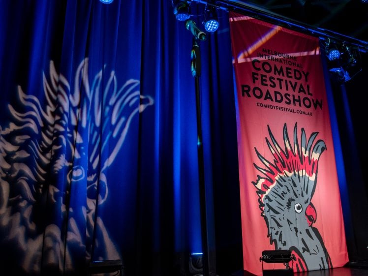 Image of A stage with blue curtains, spotlighted with an artistic bird shadow. A red banner reads “Melbourne International Comedy Festival Roadshow” with a bird illustration and the website comedfestival.com.au.