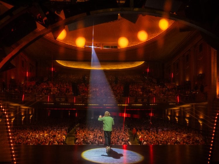 Image of A performer stands onstage under a spotlight in front of a large, packed theatre audience, with warm yellow and orange lighting illuminating the ornate auditorium.