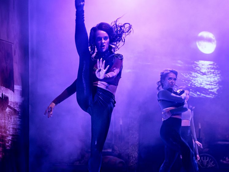 Image of A dancer performs a high kick on stage under purple lighting, with another dancer in the background. The scene features mist and a backdrop showing a moonlit seascape.