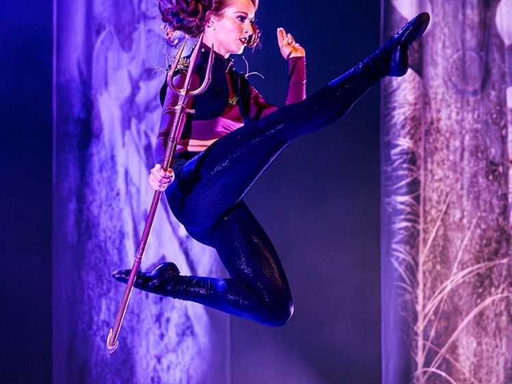 Image of A performer in a dynamic mid-air leap holds a staff, wearing a fitted costume with long sleeves and shiny leggings, against a dramatic stage backdrop with purple lighting.