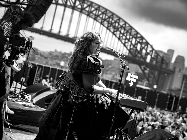 Image of A woman passionately sings and plays a keyboard on an outdoor stage with a crowd watching; the Sydney Harbour Bridge and city skyline are visible in the background under dramatic clouds.