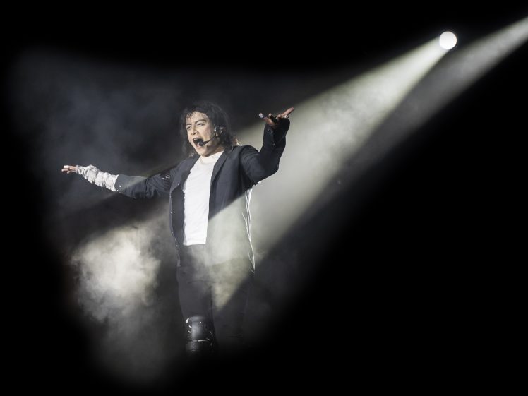 Image of A performer on stage, wearing a black jacket and white shirt, stands with arms outstretched amid smoke and dramatic spotlight beams in a dark setting.