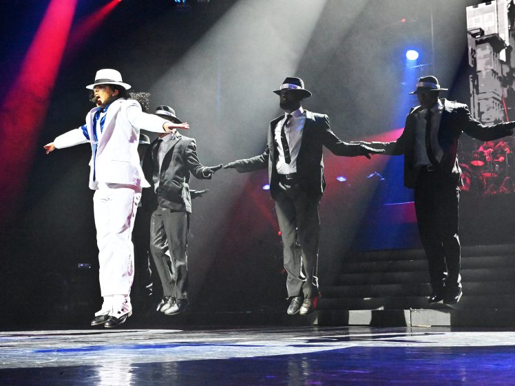 Image of Four performers in suits and hats are mid-air during a synchronised dance routine on a stage with dramatic lighting; one is dressed in white whilst the others wear dark suits.