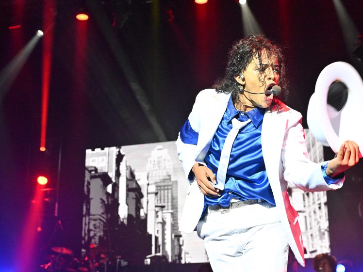 Image of A performer in a white suit with a blue shirt and tie holds a white hat and sings on stage, with dramatic lighting and a cityscape image displayed on a screen in the background.