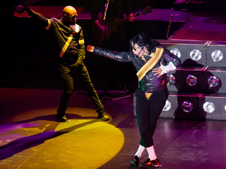 Image of A performer dressed as Michael Jackson strikes a signature dance pose on stage with a backing dancer. Both wear black and gold costumes under stage lights, with musical instruments and stairs visible in the background.