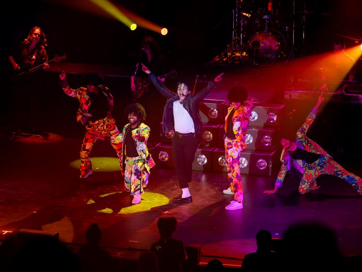 Image of A group of performers in colourful, retro outfits dance energetically on a stage with dramatic lighting, whilst a central figure dressed like Michael Jackson strikes a signature pose. Musical instruments and spotlights are visible in the background.
