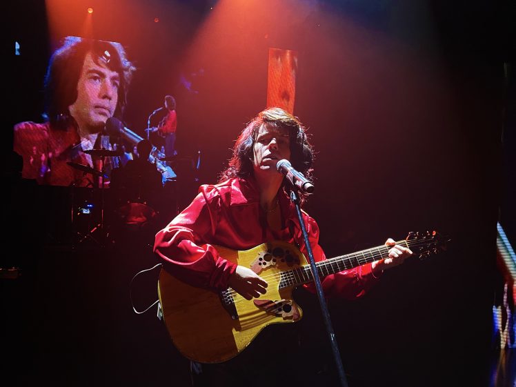 Image of A musician in a red satin shirt plays an acoustic guitar and sings into a microphone on stage, with a large screen behind showing a close-up of their face under dramatic red and orange lighting.