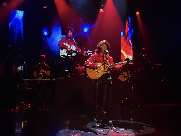Image of A musician in a red jacket plays an acoustic guitar and sings on stage under red lights, with a band in the background and a large screen showing his image behind him.