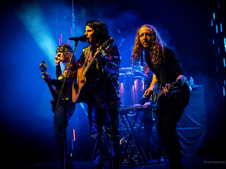 Image of Three musicians perform on a dimly lit stage; one sings into a microphone with an acoustic guitar, another plays bass, and a third plays electric guitar. Blue and purple stage lights create a vibrant atmosphere.