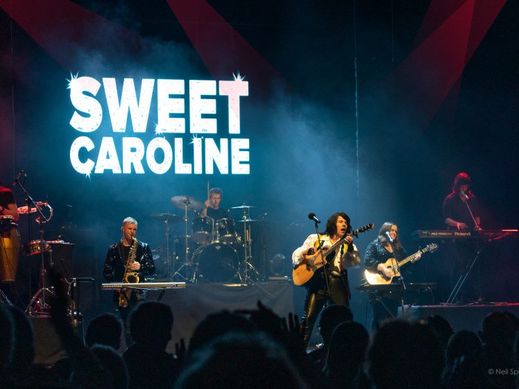 Image of A band performs on stage with bright lights, a large Sweet Caroline sign in bold white letters behind them, and several musicians playing instruments in front of an enthusiastic audience.