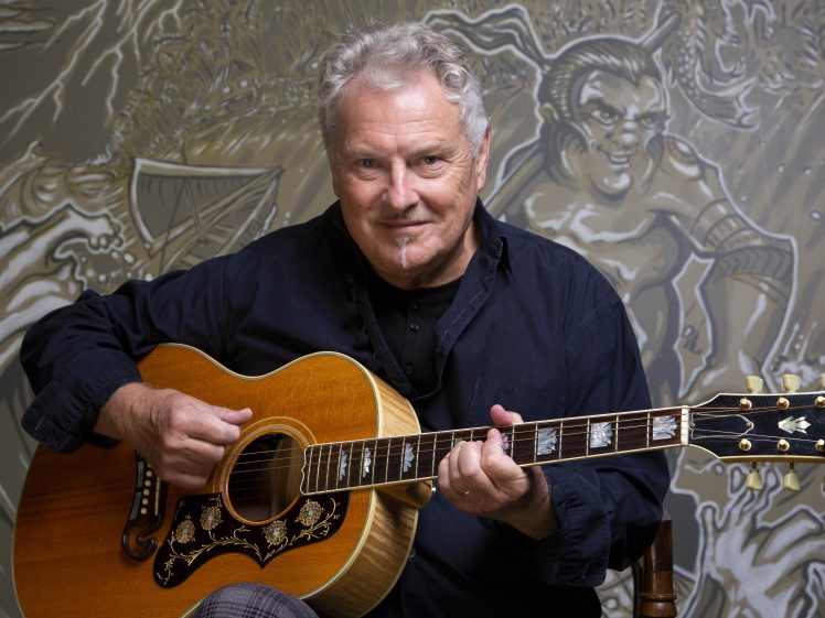 Image of An older man with grey hair sits holding an acoustic guitar, smiling at the camera. He wears a dark shirt, and there is a detailed, artistic mural with a mythical figure in the background.