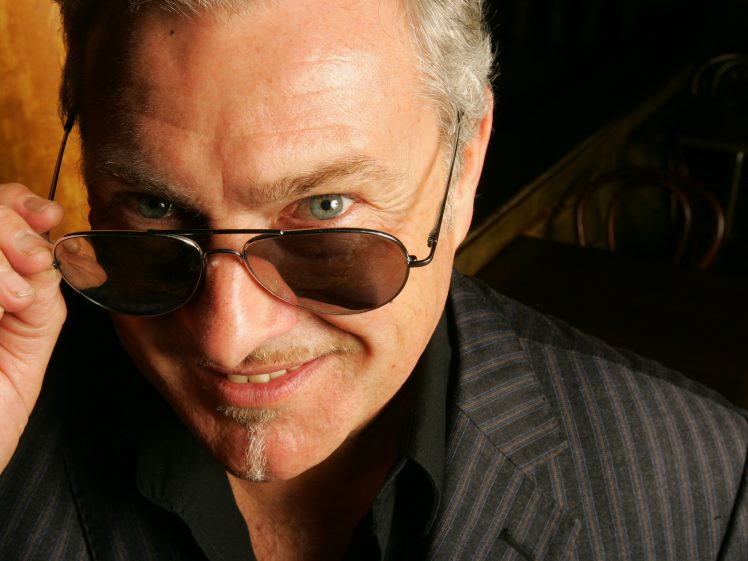 Image of A man with grey hair and a small goatee smiles whilst looking over the top of his sunglasses. He is wearing a dark pinstripe suit and is illuminated by warm lighting.