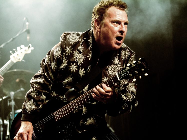 Image of A musician passionately plays an electric guitar on stage, wearing a patterned jacket. The background is dark with stage lights and some smoke, creating a dramatic atmosphere.