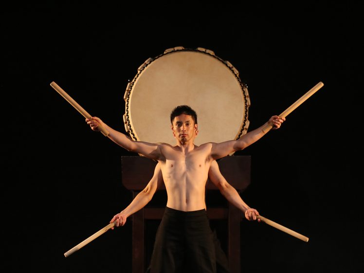 Image of A bare-chested man stands in front of a large traditional drum, holding drumsticks in each hand, with another person behind him extending their arms, creating the illusion of four arms holding drumsticks against a dark background.