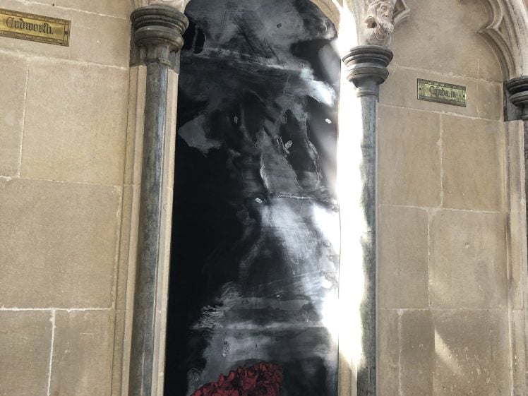 Image of A gothic stone arch with two dark marble columns frames a black panel featuring abstract white and red swirling patterns, mounted into an old stone wall with two small gold plaques on either side.