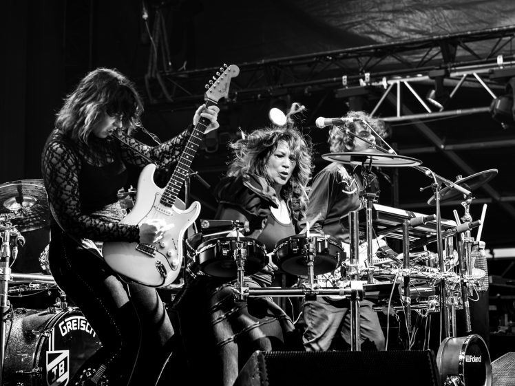 Image of Three musicians perform energetically on stage; one plays electric guitar, another plays electronic drums, and the third plays percussion. The image is in black and white, highlighting their intense expressions.