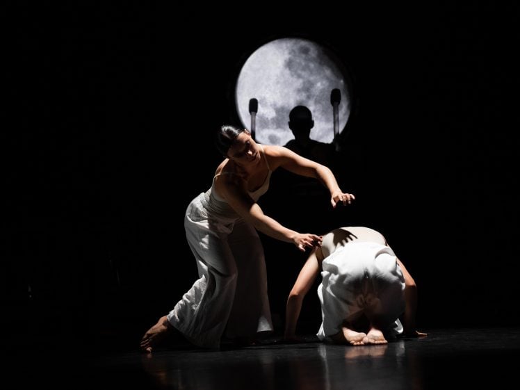 Image of Two dancers in white costumes perform on a dark stage; one is standing and gently reaching towards the other, who is kneeling and bowed. A large, illuminated moon and a shadowy figure appear in the background.