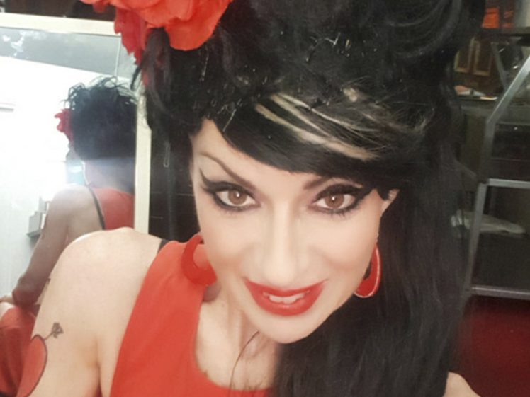 Image of A woman dressed in red with dramatic make-up, a beehive hairstyle, and a red flower hair accessory poses for a selfie. Bold white text at the top reads AMY HOUSEWINE.
