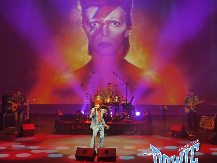 Image of A band performs on stage with a large image of David Bowie as Ziggy Stardust projected behind them. The stage is lit with colourful lights and David Bowie Major Tom text appears in the bottom right corner.